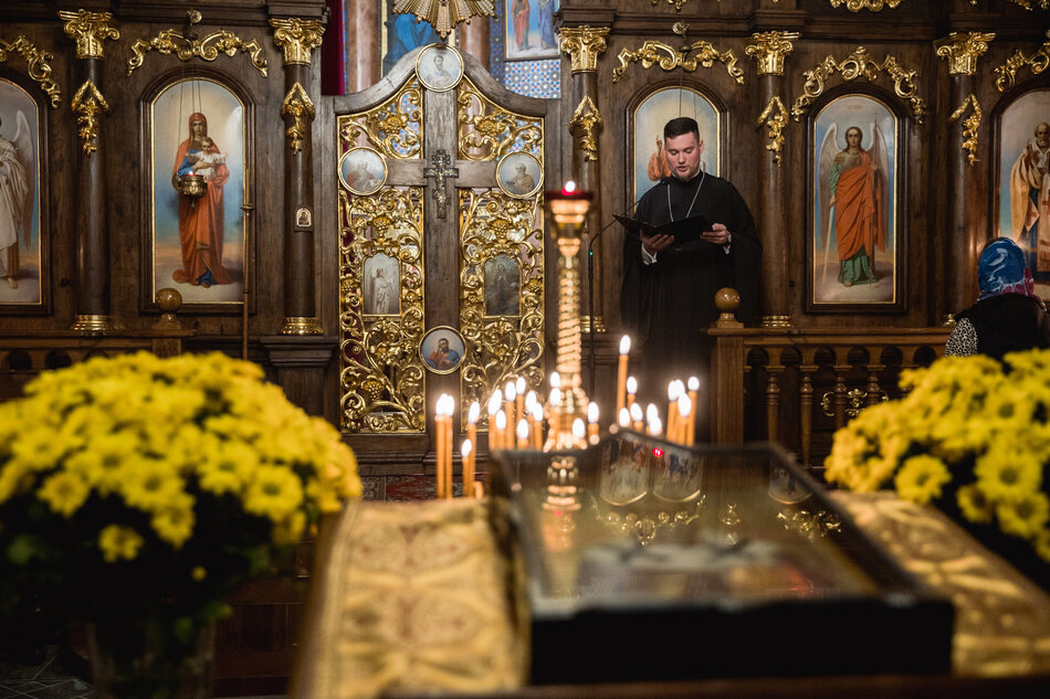 wnętrze cerkwi, widać obrazy świętych, złote zdobienia, po prawej stoi duchowny w czarnej sutannie, czyta pismo