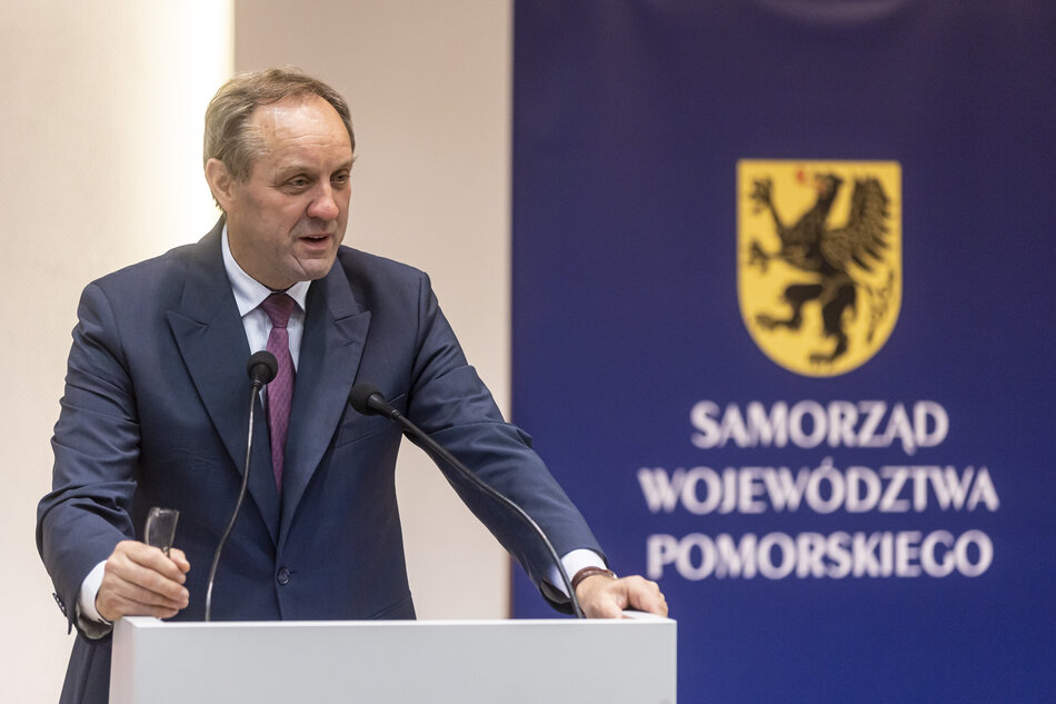 Uśmiechnięty mężczyzna po sześćdziesiątce, w popielatym garniturze, pod krawatem. Przemawia podczas konferencji. Na prawo od niego na ścianie napis Samorząd Województwa Pomorskiego i znak gryfa 