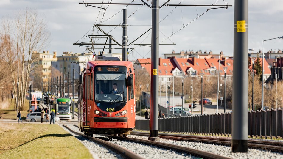 Na zdjęciu widać czerwony tramwaj o numerze bocznym 1126 poruszający się po torach. Tramwaj jest zasilany energią elektryczną przez sieć trakcyjną widoczną nad pojazdem. W tle znajdują się budynki mieszkalne z czerwonymi dachami oraz kilka innych pojazdów, w tym autobus i drugi tramwaj. Widoczna jest również infrastruktura miejska, taka jak latarnie, słupy trakcyjne oraz barierki oddzielające tory tramwajowe od chodnika. Pogoda jest słoneczna, a drzewa są bez liści, co sugeruje późną jesień lub wczesną wiosnę. Na chodniku można zauważyć kilku pieszych, w tym osoby w kamizelkach odblaskowych.