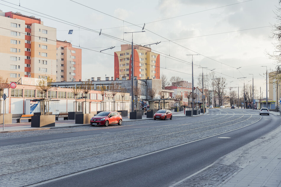 na zdjęciu jezdnia z torowiskiem tramwajowym, ulicą po prawej jedzie samochód, po lewej stoją dwa zaparkowane samochody osobowe, za nimi w tle trzy bloki mieszkaniowe