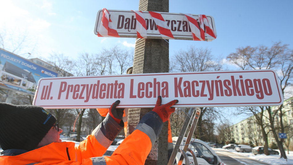 Na zdjęciu widzimy pracowników w pomarańczowych odblaskowych kurtkach, którzy montują nowy znak drogowy na słupie. Na tabliczce widnieje napis: „ul. Prezydenta Lecha Kaczyńskiego”. Nad nią znajduje się zaklejony stary znak z nazwą „Dąbrowszczaków”. Widoczny jest zimowy krajobraz w tle, z drzewami i ośnieżoną ulicą. Pracownicy używają drabiny do zamocowania tablicy, a cała scena wydaje się być częścią zmiany nazwy ulicy.