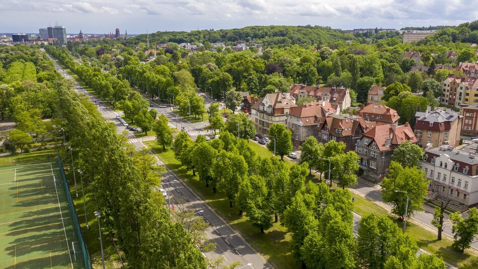 Na zdjęciu widzimy panoramę miejskiej dzielnicy, prawdopodobnie w Polsce. Przez centralną część obrazu przepływa rzeka, obramowana zielenią oraz alejkami spacerowymi. Wzdłuż rzeki znajdują się budynki o zróżnicowanej architekturze – od starszych kamienic po nowoczesne obiekty. W dolnej części obrazu widoczny jest zielony teren rekreacyjny z alejkami, ławkami i placem zabaw. W tle rozciąga się rozbudowana część miasta, z wieloma budynkami mieszkalnymi i przemysłowymi, a także z widocznymi mostami. W dalszej perspektywie można dostrzec obszary zielone oraz bardziej odległe struktury miejskie. Jest to widok typowy dla miejskiego krajobrazu zharmonizowanego z naturalnym otoczeniem.