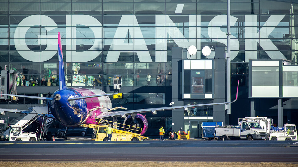 Na zdjęciu widzimy lotnisko w Gdańsku, co można rozpoznać po wielkim napisie GDAŃSK na fasadzie terminalu. Na pierwszym planie znajduje się samolot w kolorach różowo-niebieskich linii lotniczych Wizz Air. Wokół samolotu trwają operacje obsługowe – widoczne są pojazdy techniczne, takie jak schody, pojazdy do transportu bagażu i obsługi naziemnej, a także personel techniczny w odblaskowych kamizelkach. W tle za szybami terminalu można dostrzec wnętrze budynku oraz pasażerów i dekoracje. Scena przedstawia typowy obrazek z ruchliwego lotniska, gdzie przygotowania do lotów odbywają się w pełnym biegu.