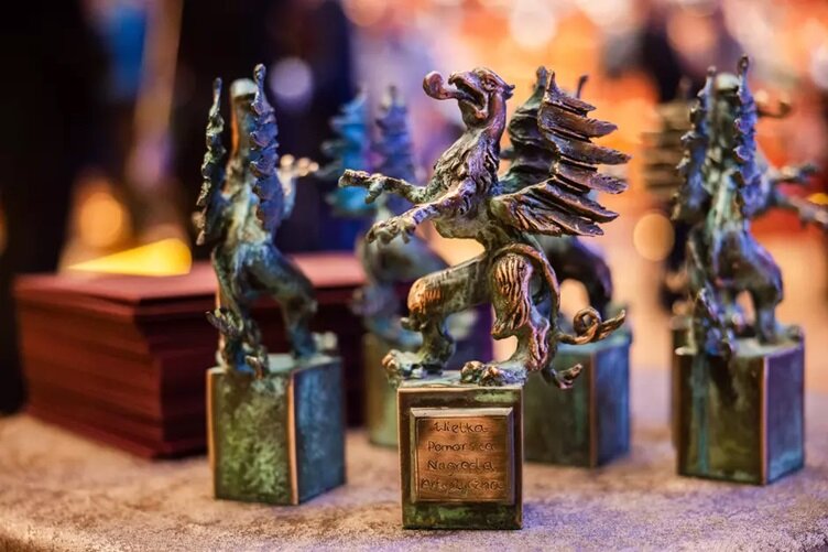 Zdjęcie przedstawia brązowe statuetki w kształcie gryfów, które są nagrodami artystycznymi. Każda statuetka umieszczona jest na podstawie z wygrawerowaną nazwą Pomorska Nagroda Artystyczna, a w tle widać rozmazane światła, nadające uroczysty klimat