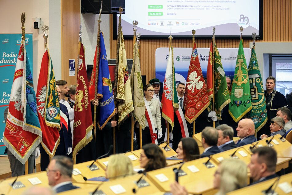 W dużej sali konferencyjnej stoi kilkanaście osób ze sztandarami przed ludźmi siedzącymi na krzesłach.