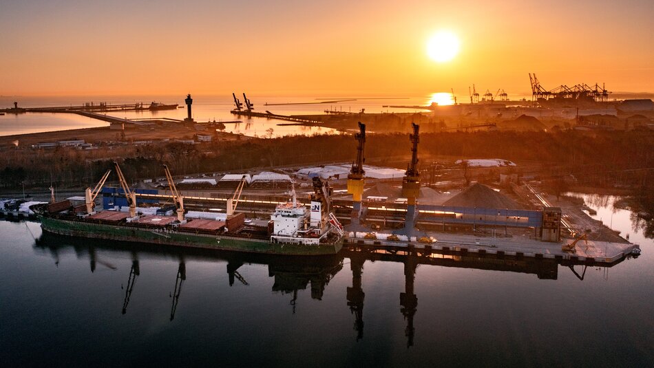 Na zdjęciu widać port morski o poranku lub podczas zachodu słońca, co podkreśla piękne, ciepłe światło słoneczne odbijające się w wodzie. Na pierwszym planie znajduje się duży statek towarowy przy nabrzeżu, wyposażonym w dźwigi portowe służące do załadunku i rozładunku towarów. W tle widać rozległą infrastrukturę portową, z magazynami, terminalami i innymi statkami. Całość jest otoczona wodą oraz terenami leśnymi i przemysłowymi. Krajobraz emanuje spokojem, a zarazem wskazuje na intensywną działalność przemysłową. To miejsce prawdopodobnie znajduje się w Gdańsku, w porcie, który jest jednym z najważniejszych w Polsce.