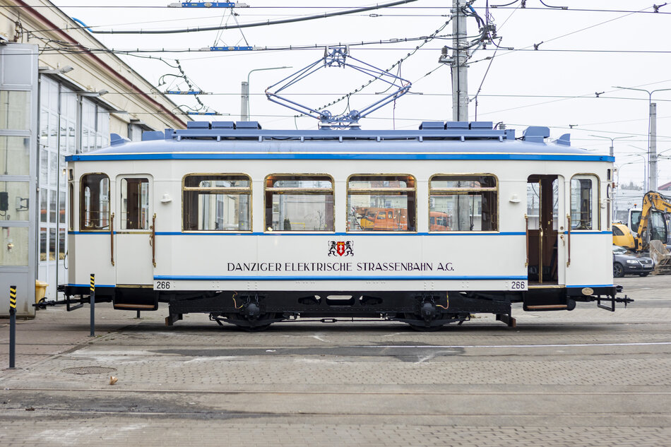 Zdjęcie przedstawia zabytkowy tramwaj „Danziger Elektrische Strassenbahn A.G.” oznaczony numerem 266, stojący na terenie zajezdni tramwajowej. Tramwaj wyróżnia się eleganckim malowaniem w kolorze białym z niebieskimi akcentami oraz detalami odzwierciedlającymi styl z epoki, w której powstał.