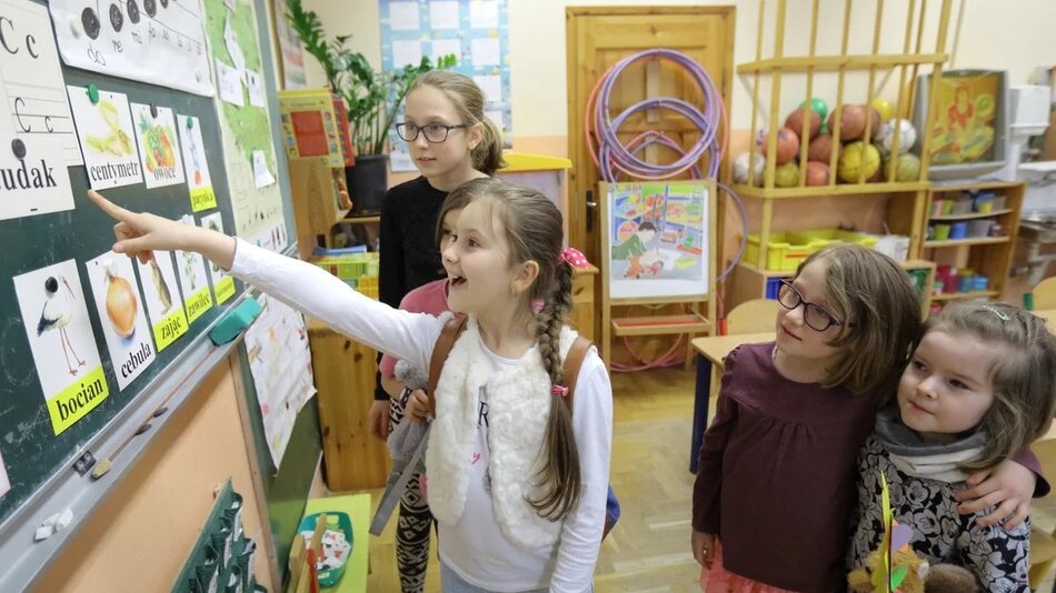 Nauczycielka i troje dzieci stoi w klasie przed tablicą, na której przypięte są rysunki
