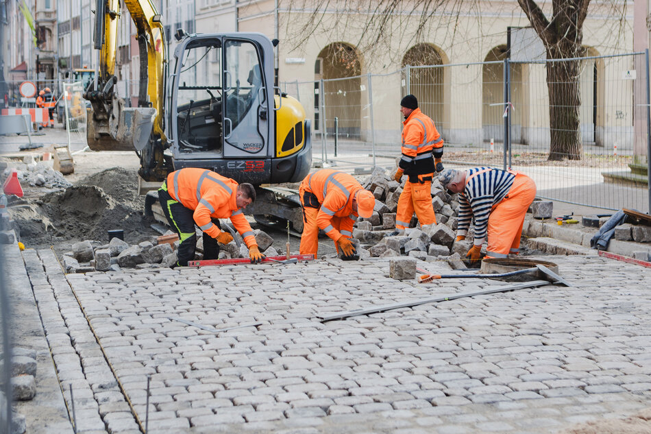 na zdjęciu czterech robotników w pomarańczowych kombinezonach, układają kostkę brukową na ulicy