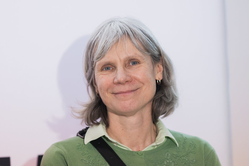 W zbliżeniu do kamery uśmiecha się kobieta w średnim wieku z krótkimi siwymi włosami i niebieskimi oczami. Nosi jasnoniebieską koszulkę z białym kołnierzem. Koszula ma czarny pasek na przodzie, który jest odlewany na ścianie za nią, dodając odrobinę głębi scenie