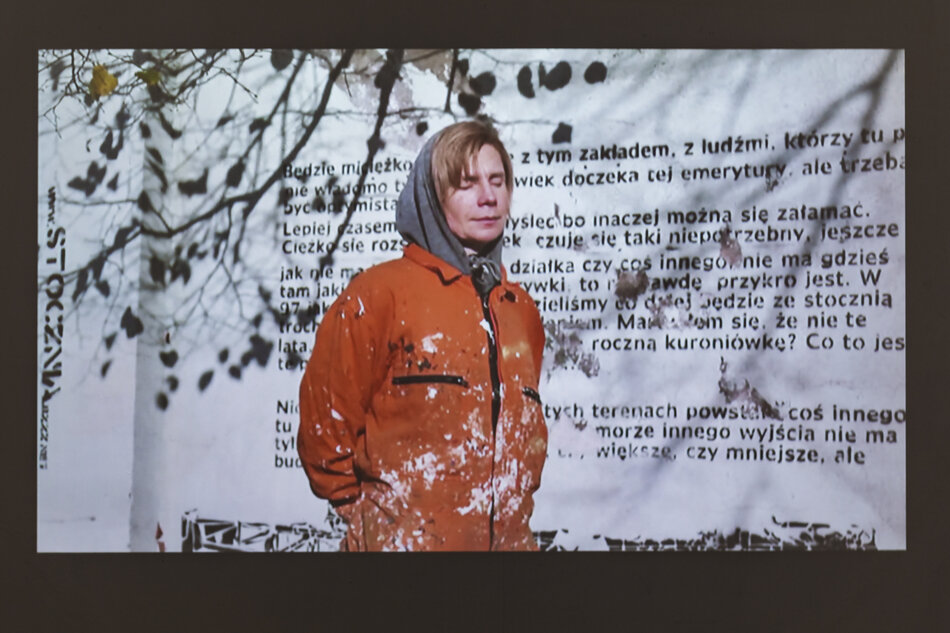 Kobieta w pomarańczowym kombinezonie roboczym stoi na tle muru. Ma zamknięte oczy