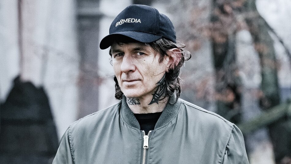 Mężczyzna w zielonej kurtce i czarnej czapce z daszkiem. Mężczyzna ma dłuższe ciemne włosy i tatuaże na szyi.