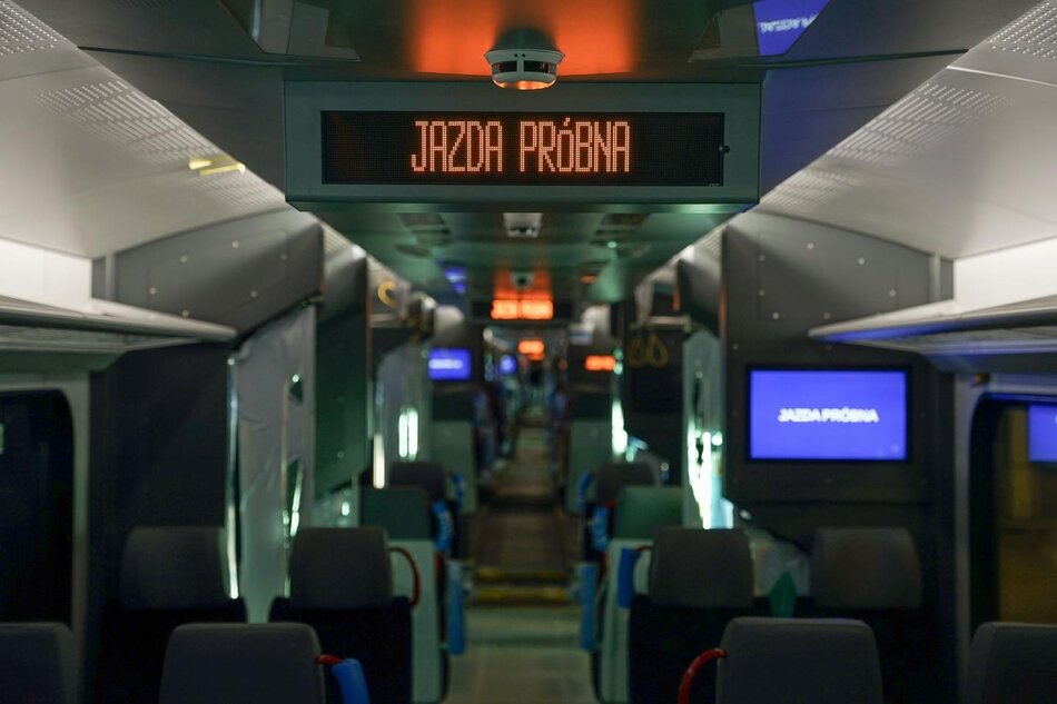 Zdjęcie przedstawia wnętrze nowoczesnego pociągu z wyświetlaczem informującym o JAZDIE PRÓŻNEJ, co oznacza, że pojazd jest na testowym lub technicznym przejeździe bez pasażerów. Wnętrze jest dobrze oświetlone, z nowoczesnymi siedzeniami i ekranami informacyjnymi, co sugeruje wysoki standard podróży.