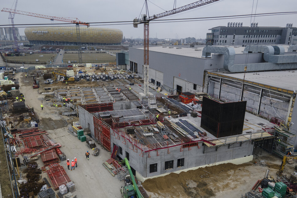 Na zdjęciu widoczny jest plac budowy nowego obiektu w pobliżu hali Amber Expo w Gdańsku. W tle znajduje się charakterystyczny, złoty budynek stadionu Polsat Plus Arena Gdańsk. W centralnej części fotografii widać konstrukcję nowego budynku, wykonaną częściowo z betonu, z elementami metalowymi oraz drewnianymi. Prace budowlane są w zaawansowanym stadium – na miejscu pracują robotnicy w kaskach i odblaskowych kamizelkach, a w otoczeniu znajdują się liczne materiały budowlane, stalowe rusztowania i maszyny. Dźwig budowlany góruje nad placem budowy, co sugeruje, że konstrukcja nadal będzie wznoszona na większą wysokość. Po prawej stronie widać duży szary budynek z wentylacją, co może wskazywać na istniejący już obiekt przemysłowy lub magazynowy. Na placu budowy znajduje się wiele pojazdów, w tym koparki i ciężarówki, a także rzędy betonowych prefabrykatów.