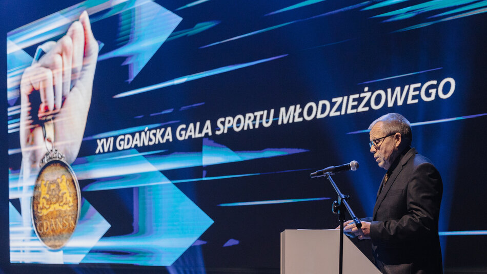 Na zdjęciu widać mężczyznę przemawiającego przy mównicy podczas uroczystości na tle ekranu z napisem XVI Gdańska Gala Sportu Młodzieżowego. Na ekranie widoczna jest również grafika przedstawiająca rękę trzymającą medal z herbem Gdańska.
