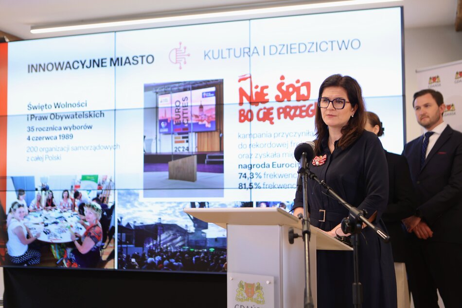 kobieta w czarnej sukience stoi za pulpitem, mówi do mikrofonu, w tle po prawej stoją dwie osoby