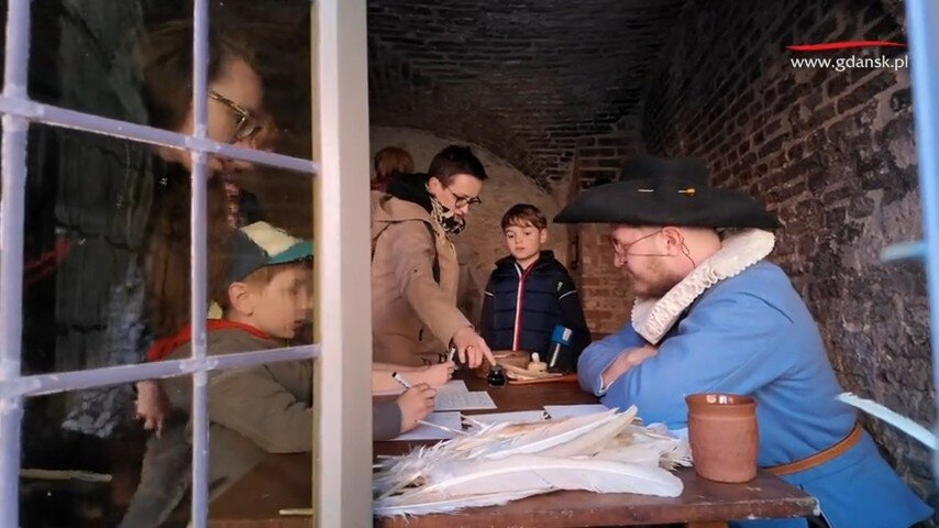 Na zdjęciu widzimy grupę dzieci i dorosłych uczestniczących w edukacyjnej inscenizacji historycznej – piszą oni gęsim piórem przy drewnianym stole, naprzeciwko mężczyzny w historycznym stroju z epoki. Cała scena odbywa się w kamiennej izbie, a przez szybę widać odbicie dodatkowych osób oraz adres strony internetowej miasta Gdańsk.