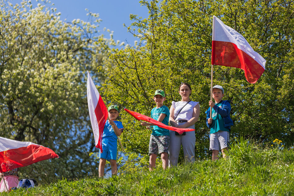 Kobieta stoi na zielonym od trawy wzgórzu, po jej obu stronach są mali chłopcy z biało-czerwonymi flagami 