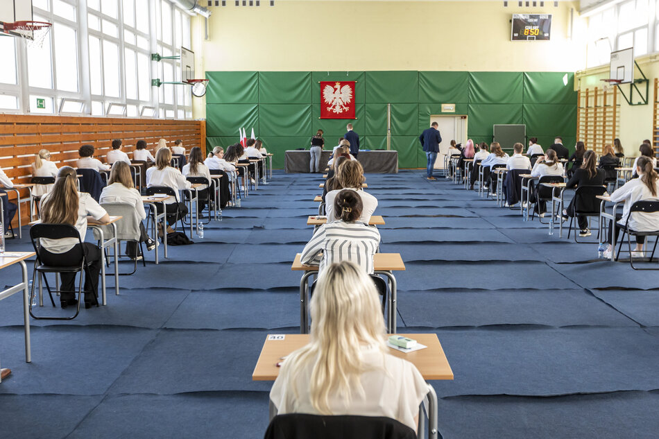 Na zdjęciu widać salę gimnastyczną przekształconą w miejsce egzaminacyjne. Uczniowie siedzą pojedynczo przy oddzielnych stolikach, rozmieszczonych w równych odstępach, co sugeruje, że odbywa się ważny egzamin, prawdopodobnie maturalny. Wszyscy są ubrani elegancko, głównie na biało-czarno, co jest typowe dla oficjalnych okazji szkolnych w Polsce. Na ścianie z przodu sali widnieje godło Polski – biały orzeł na czerwonym tle – co dodatkowo podkreśla oficjalny charakter wydarzenia. Obok znajduje się stół komisji egzaminacyjnej oraz flaga Polski. Na zegarze w rogu sali widnieje godzina 8:50. Cała scena wygląda na bardzo uporządkowaną i formalną, zgodnie z zasadami przeprowadzania egzaminów państwowych w Polsce.
