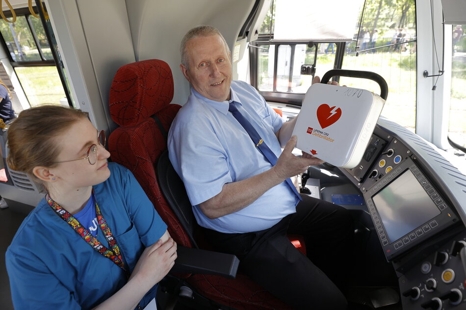 Na zdjęciu widzimy kierowcę autobusu siedzącego w kabinie, który trzyma w rękach defibrylator z widocznym symbolem serca. Obok niego siedzi młoda kobieta w medycznym uniformie, prawdopodobnie ratowniczka lub pielęgniarka, która z zainteresowaniem patrzy na urządzenie.