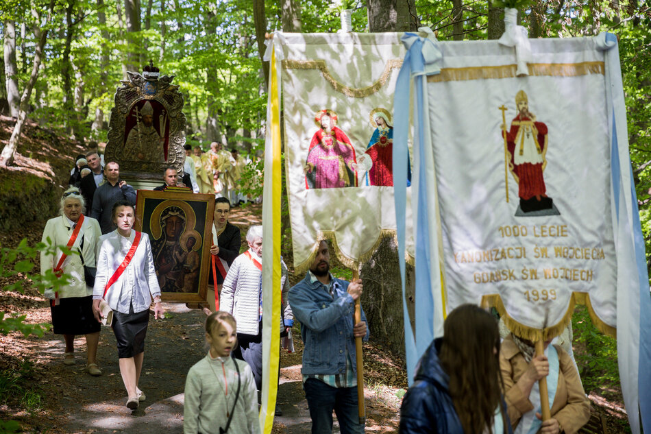  Grupa ludzi idących ścieżką w lesie, noszą transparenty z obrazami religijnymi i tekstem na nich, banery wydają się być związane z procesją religijną lub procesją, niektórzy ludzie noszą czerwono-białe szaszły, wskazujące, że są częścią większej grupy, w tle są drzewa i krzewy, sugerujące, iż zdjęcie zostało zrobione w ciągu dnia, w środku obrazu możemy zobaczyć duży posąg Jezusa Chrystusa, z koroną cierniową na górze i transparentem z napisem 1000 Lecie (1000 lat służby) nad nim, po prawej stronie tego transparentu jest również ozdobiony obrazami świętych i innych postaci religijnych, poniżej posągu znajduje się mniejszy transparent z tekstem i liczbami, prawdopodobnie upamiętniający 1000-lecie służenia św. Jana Chrzciciela, jest też kilkoro dzieci i dorosłych