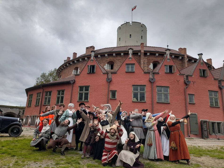 Na zdjęciu grupa ludzi w strojach historycznych pozuje w radosny i teatralny sposób na tle czerwonego budynku z wieżą i polską flagą, co sugeruje udział w rekonstrukcji historycznej lub wydarzeniu tematycznym. Sceneria i architektura wskazują na historyczne miejsce, prawdopodobnie zlokalizowane w Polsce.
