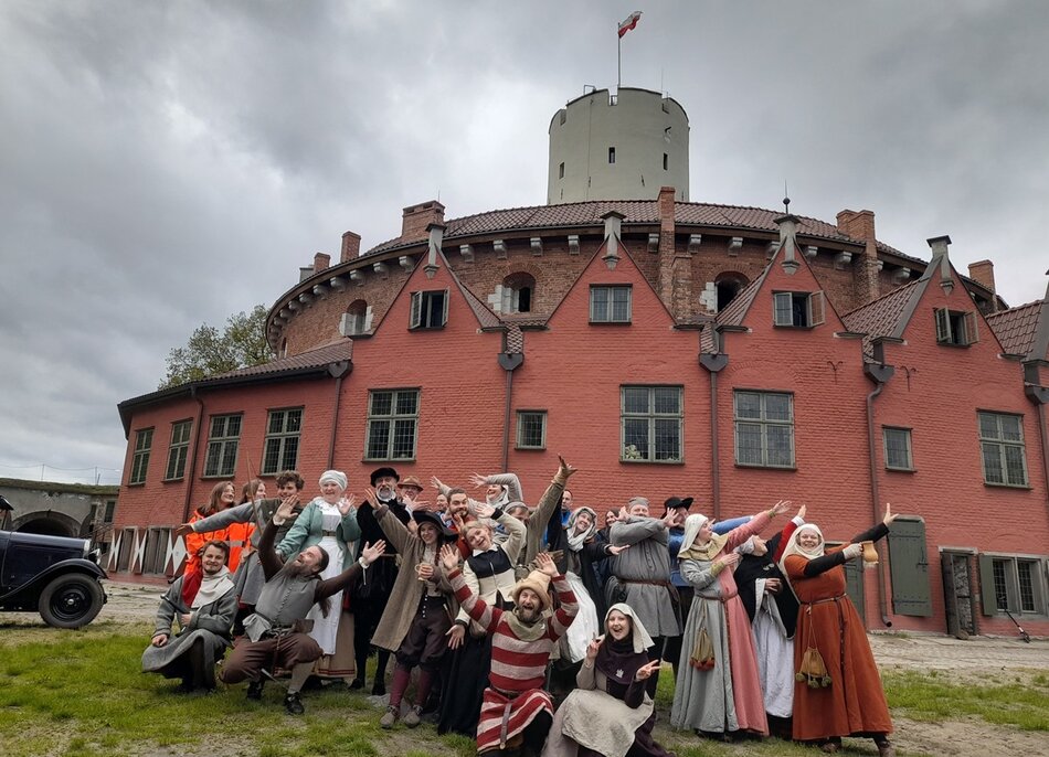 Na zdjęciu grupa ludzi w strojach historycznych pozuje w radosny i teatralny sposób na tle czerwonego budynku z wieżą i polską flagą, co sugeruje udział w rekonstrukcji historycznej lub wydarzeniu tematycznym. Sceneria i architektura wskazują na historyczne miejsce, prawdopodobnie zlokalizowane w Polsce.