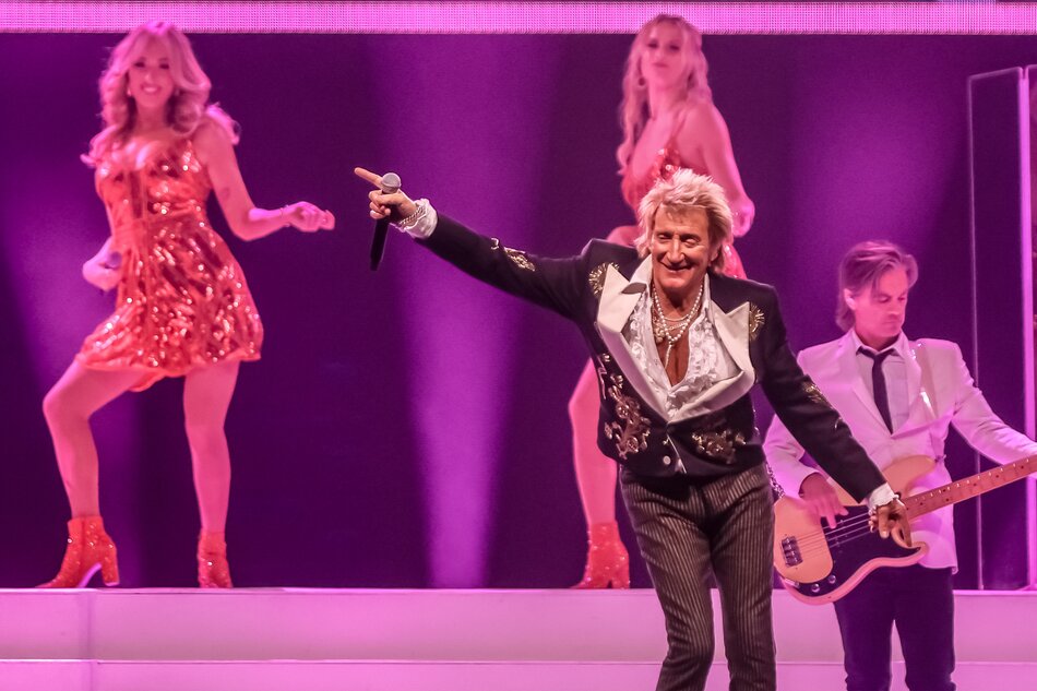 na zdjęciu rod stewart tańczący na scenie, za nim dwie tancerki i gitarzysta