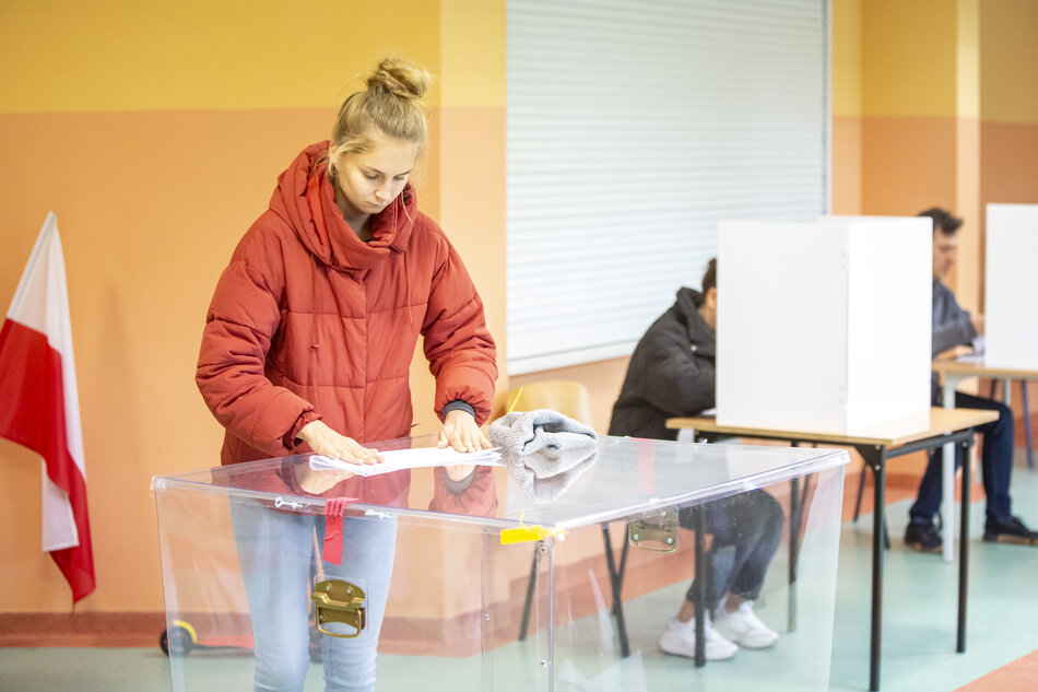  młoda kobieta w czerwonej kurtce stoi przed przezroczystym pudełkiem wyborczym, trzyma w ręku kartkę papieru i wydaje się oddawać głos, pudełko umieszczone jest na stole z żółtą wstążką związaną wokół niego, w tle są inni ludzie siedząc przy stołach z stoiskami do głosowania, a flaga Polski po lewej stronie znajduje się na stacji wyborczej.
