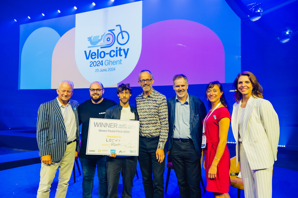 Na zdjęciu widzimy grupę siedmiorga uśmiechniętych osób stojących na scenie podczas wydarzenia Velo-city 2024 w Gandawie, które odbyło się 20 czerwca 2024 roku. W centrum znajduje się zwycięzca konkursu „Smart Pedal Pitch 2024”, trzymający dużą tablicę z napisem „WINNER” i nazwą projektu „LOCKY”.