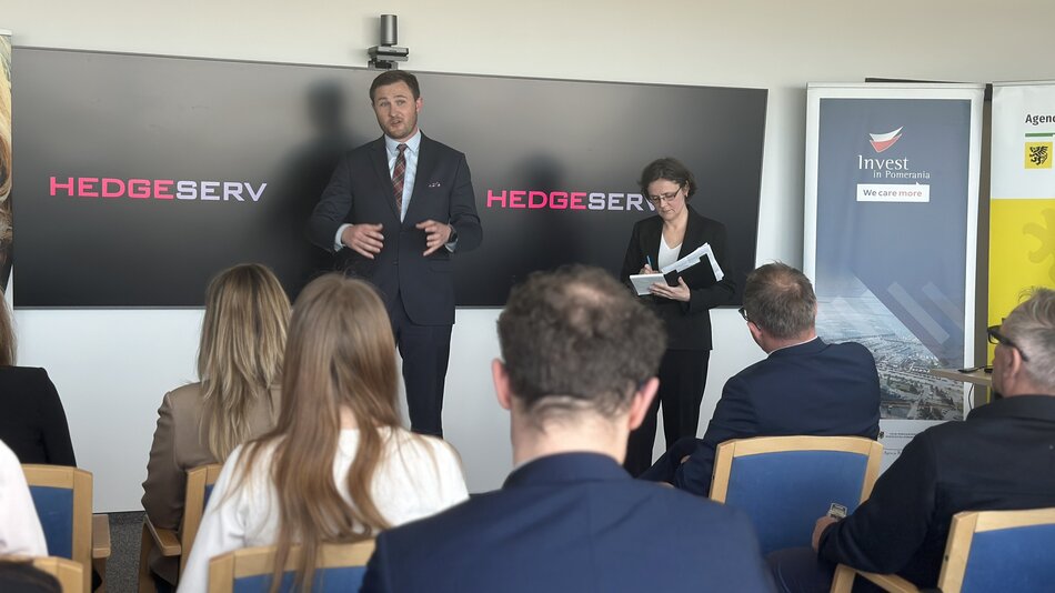Na zdjęciu widzimy wydarzenie biznesowe lub konferencję, w której bierze udział grupa ludzi. Na pierwszym planie znajduje się publiczność siedząca tyłem do obiektywu, słuchająca prezentacji. Na tle dwóch dużych ekranów z logo firmy „HEDGESERV” stoi mężczyzna w garniturze, który najwyraźniej prowadzi prezentację lub przemówienie. Obok niego znajduje się kobieta w garniturze, notująca coś w zeszycie, co może sugerować, że pełni rolę asystentki, dziennikarki lub osoby wspierającej prezentację.