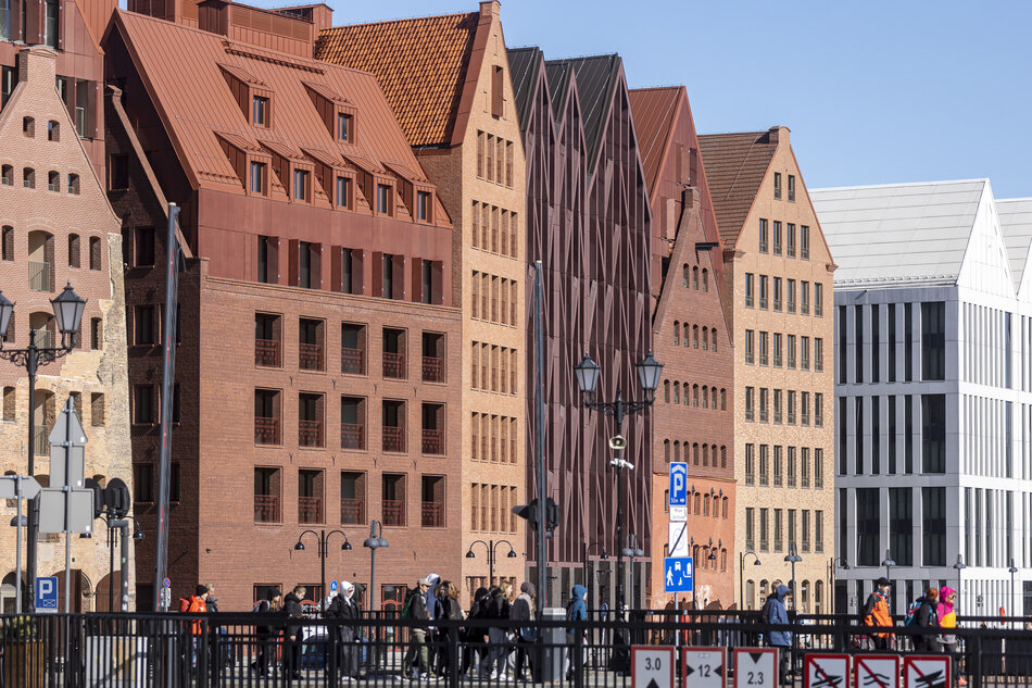 rząd wielopiętrowych budynków na rogu ulicy. Budynki są wykonane z cegły i mają czerwony dach płytkowy. Ułożone są w linii przekątnej, z najwyższym budynkiem po lewej i najkrótszym budynku pośrodku. Po prawej stronie jest wysoki biały budynek z zakrzywionym dachem i szklaną fasadą. Przed każdym budynkami widać grupę ludzi idących ulicą, a na całej scenie rozrzucone są lampy i znaki drogowe.