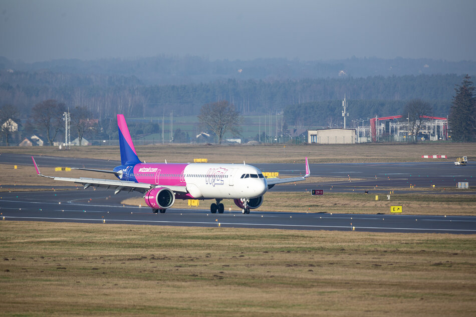 Na zdjęciu widoczny jest samolot linii Wizz Air kołujący po pasie startowym, otoczony rozległą przestrzenią lotniska i zielonymi terenami w tle. Charakterystyczne różowo-fioletowe barwy maszyny kontrastują z naturalnym krajobrazem, podkreślając dynamikę współczesnego transportu lotniczego.