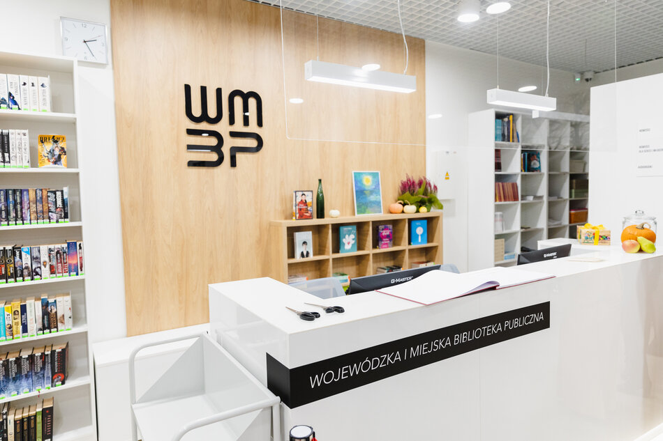Zdjęcie przedstawia nowoczesne wnętrze biblioteki publicznej o nazwie „Wojewódzka i Miejska Biblioteka Publiczna”, z jasnym wystrojem, ladą recepcyjną i estetycznie rozmieszczonymi książkami. Na ścianie za ladą widnieje czarne logo „WMBP”, a obok znajdują się dekoracje, owoce i regał z literaturą i ozdobami