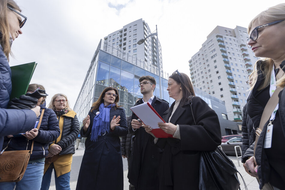 grupka osób stoi w okręgu, rozmawiają, w tle wysokie bloki