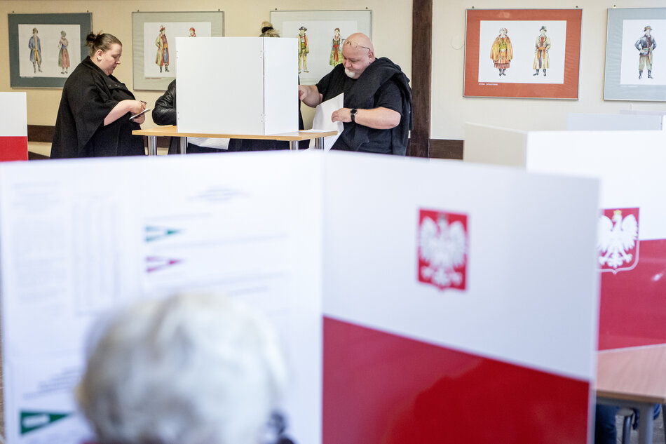 Na zdjęciu widzimy ludzi głosujących w lokalu wyborczym. Scena ma miejsce w Polsce, o czym świadczą biało-czerwone parawany z godłem państwowym (orłem w koronie). Kilka osób wypełnia karty do głosowania przy stanowiskach zasłoniętych dla zachowania prywatności. W tle widoczne są ilustracje przedstawiające postacie w tradycyjnych, historycznych strojach polskich. Atmosfera jest spokojna, a ludzie skupieni na oddaniu głosu.