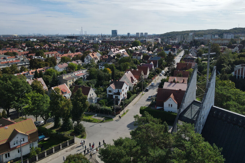 zdjęcie z drona, widać dzielnicę pełną domów jednorodzinnych ustawionych w kilku rzędach, pomiędzy nimi dużo zielonych drzew, w tle kilka drapaczy chmur