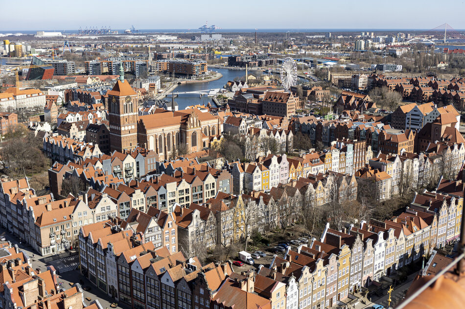 Zdjęcie przedstawia panoramę Gdańska, widzianą z wieży kościoła Mariackiego, z charakterystycznymi kolorowymi kamienicami, gotycką bazyliką oraz nowoczesnymi budynkami i kołem widokowym w tle. W oddali widać port morski i nabrzeże, co podkreśla nadmorskie położenie miasta. 