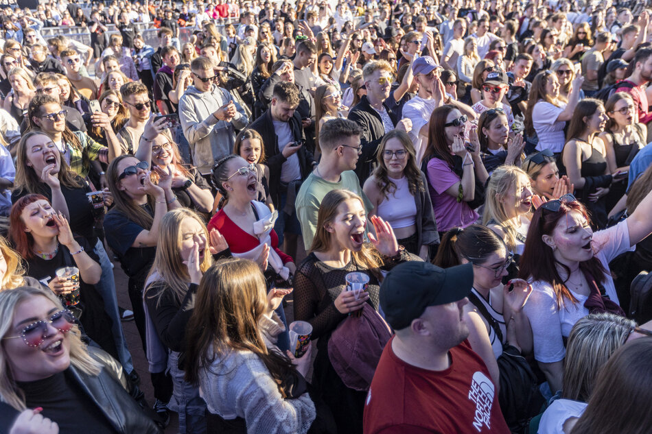  Duży tłum ludzi zebranych na festiwalu muzycznym. Tłum składa się zarówno z mężczyzn, jak i kobiet, a wszyscy uśmiechają się i dopingują rękami w powietrzu. Niektórzy z nich trzymają telefony, aby robić zdjęcia lub filmy, podczas gdy inni patrzą w niebo. W tle są drzewa i budynki, sugerujące, że festiwal odbywa się na zewnątrz.