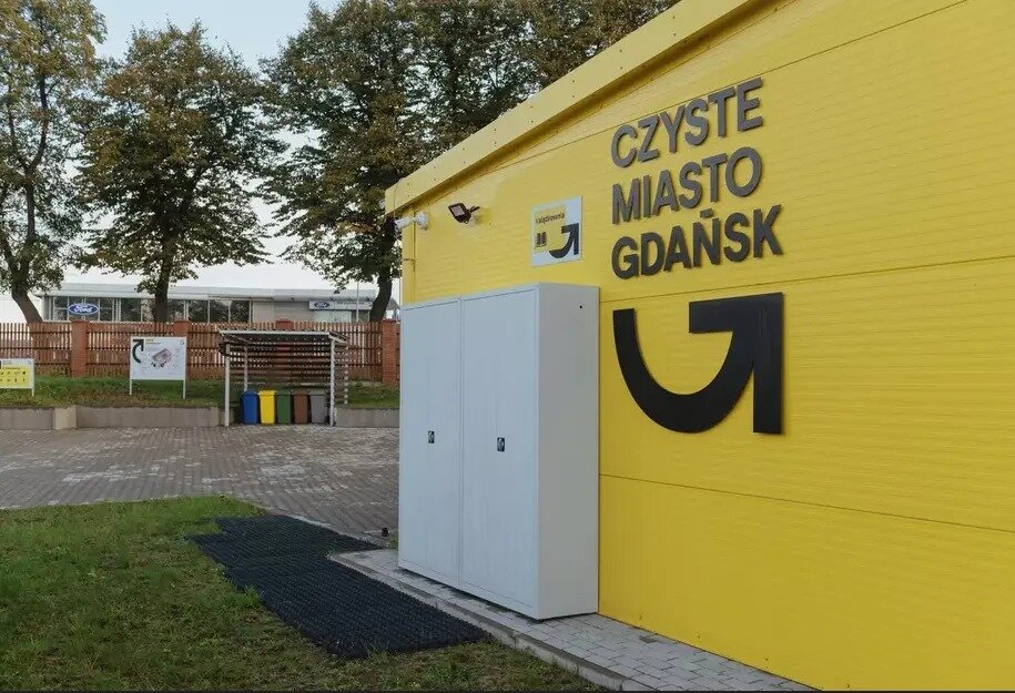 Na zdjęciu widoczny jest jasnożółty budynek z napisem „Czyste Miasto Gdańsk” oraz charakterystycznym logotypem programu gospodarki odpadami. W tle znajduje się uporządkowany teren z pojemnikami na odpady i tablicami informacyjnymi, co sugeruje, że to punkt selektywnej zbiórki odpadów komunalnych (PSZOK). 