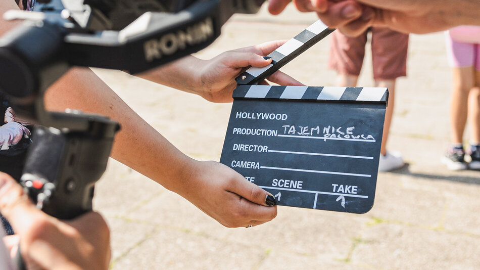 Na zdjęciu widzimy klaps filmowy używany podczas kręcenia filmu. Jest on trzymany przez dwie osoby, a na górze klapsa widnieje napis „HOLLYWOOD”. Poniżej w polu „PRODUCTION” odręcznie zapisano tytuł: „TAJEMNICE FALOWCA”. Inne pola, takie jak „DIRECTOR” czy „CAMERA”, są puste. W polu „TAKE” wpisano cyfrę „1”.