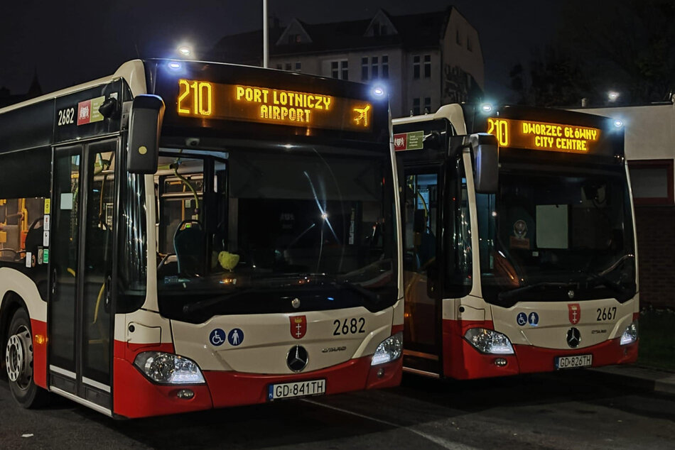 Dwa autobusy stoją zaparkowane. Na ich wyświetlaczach widać komunikaty po angielsku i ikonę samolotu. Zdjęcie zrobione noca.