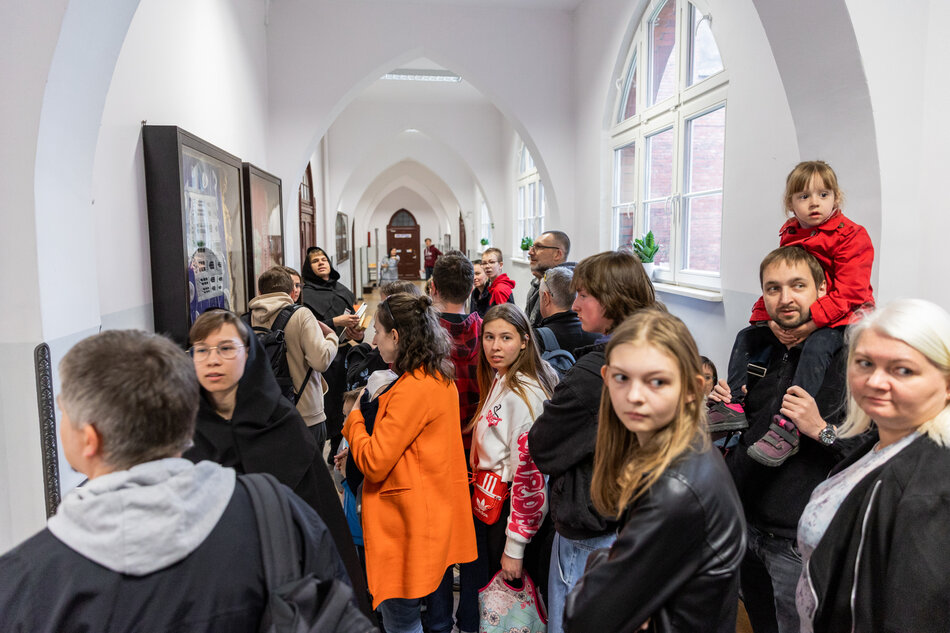 Tłum ludzi znajduje się w jasnym, zabytkowym korytarzu z gotyckimi sklepieniami, najwyraźniej czekając na wejście do jednego z pomieszczeń muzealnych. Wśród odwiedzających są osoby w różnym wieku, w tym dzieci, co świadczy o rodzinnej atmosferze wydarzenia