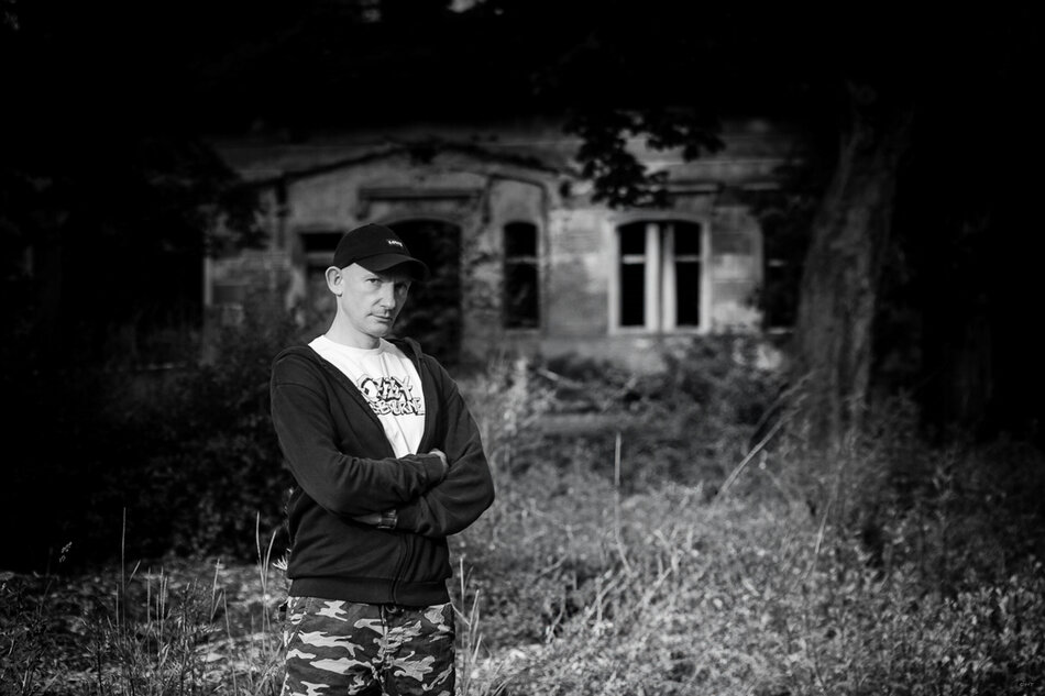 Na zdjęciu mężczyzna z założonymi rękoma, w ciemnej bluzie i czapeczce z daszkiem, w tle stary dom.
