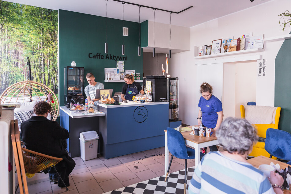 Zdjęcie przedstawia przytulną kawiarnię „Caffe Aktywni”, w której kilka osób pracuje za ladą i obsługuje gości, a inni relaksują się przy stolikach. Wnętrze wyróżnia się ciepłą atmosferą, zieloną ścianą z leśnym motywem i licznymi certyfikatami na półce, co podkreśla zaangażowanie społeczności w rozwój tego miejsca. 