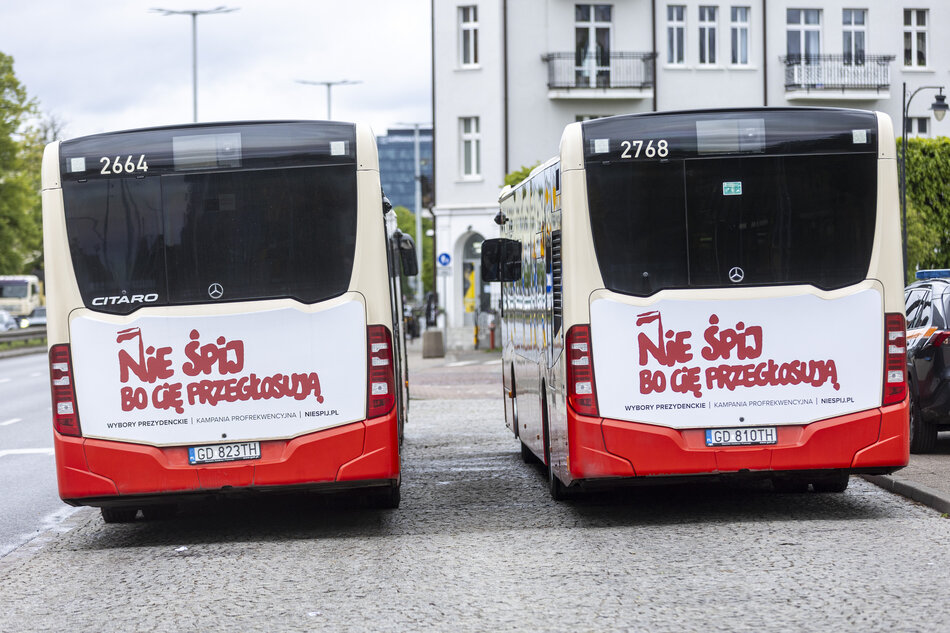 Dwa autobusy stoją zaparkowane na przystanku w mieście, widać je od tyłu. Na autobusach widać hasło namalowane czerwoną czcionką solidarycą: Nie śpij bo cię przegłosują.