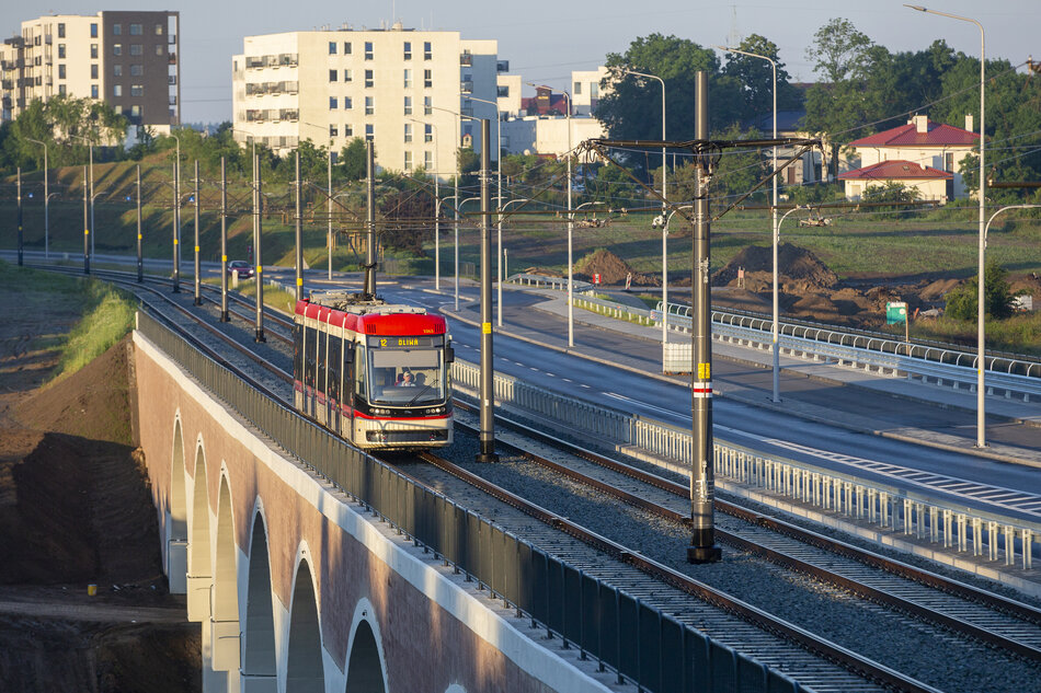 Na zdjęciu widoczny jest tramwaj linii 12 jadący po nowoczesnym wiadukcie w słoneczny poranek, z osiedlem mieszkaniowym i zielenią w tle. 