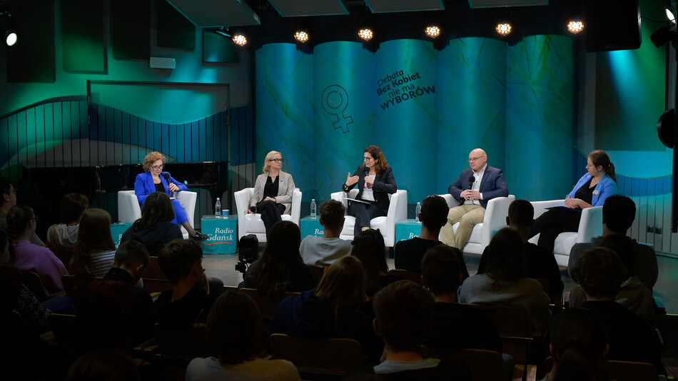 Na zdjęciu widzimy debatę publiczną zorganizowaną przez Radio Gdańsk. Wydarzenie odbywa się na scenie z profesjonalnym oświetleniem i tłem w kolorze niebiesko-zielonym, na którym widnieje hasło: „Debata. Bez kobiet nie ma wyborów” oraz symbol kobiety. Na scenie siedzi pięcioro panelistów – cztery kobiety i jeden mężczyzna – na białych fotelach, z mikrofonami w rękach. Przed nimi siedzi widownia, głównie młodzi ludzie. Paneliści wyglądają na zaangażowanych w dyskusję – jedna z kobiet właśnie mówi, reszta słucha lub przegląda notatki.