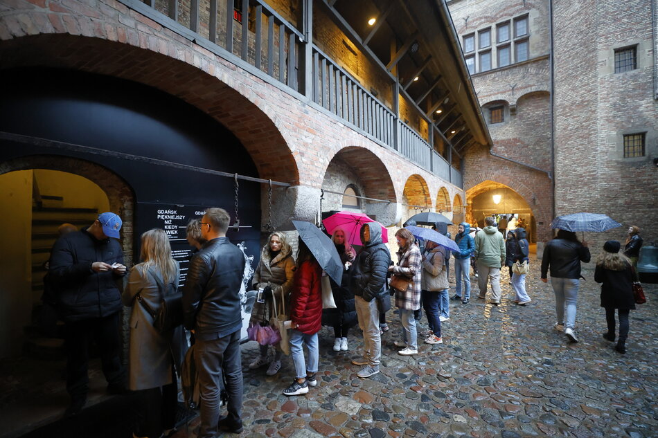Ludzie z parasolami czekają w długiej kolejce przed wejściem do muzeum zlokalizowanego w zabytkowej kamienicy z arkadami. Mimo deszczu panuje ożywiona atmosfera, co świadczy o dużym zainteresowaniu wydarzeniem