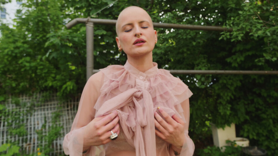 Na zdjęciu kobieta bez włosów, w sukni z delikatnego materiału, na palcu duży czarny pierścień, w tle zieleń i trzepak.